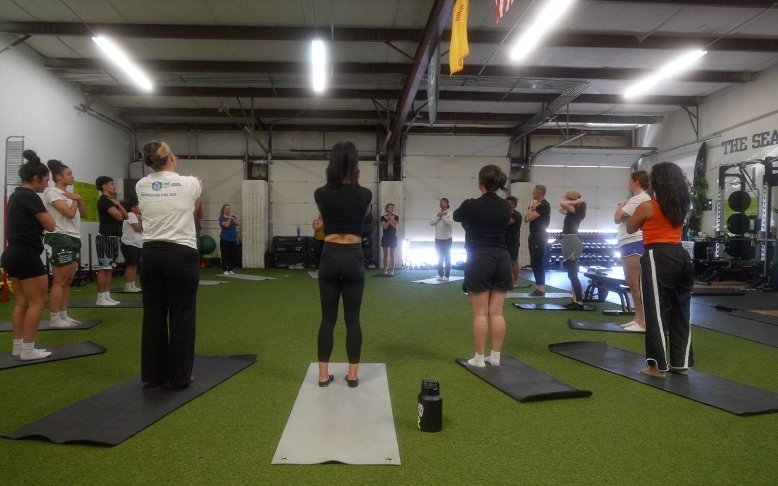 A room of people standing on mats, arms crossed on the chest, mid breath